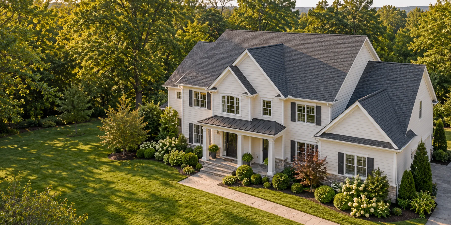 White home with a charcoal-shingle roof, mature trees and landscaped lawn